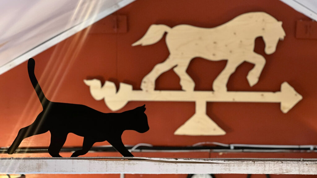 black cat cut out in front of a wooden horse-shaped weather vane on a red barn background
